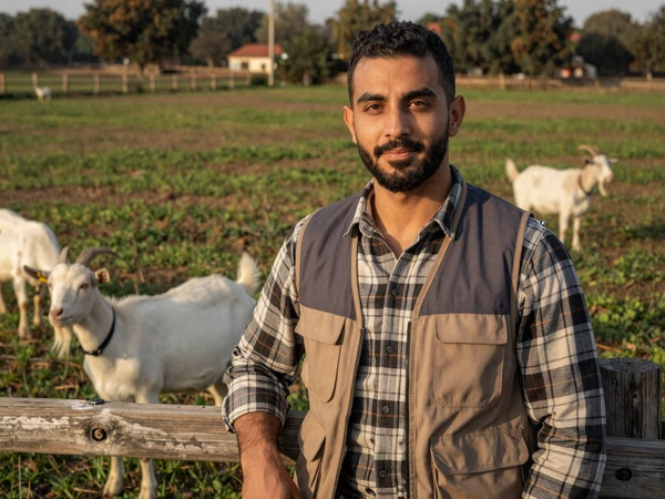 Ibrahim, a Muslim American farmer standing near a pasture fence with goats grazing in warm afternoon light